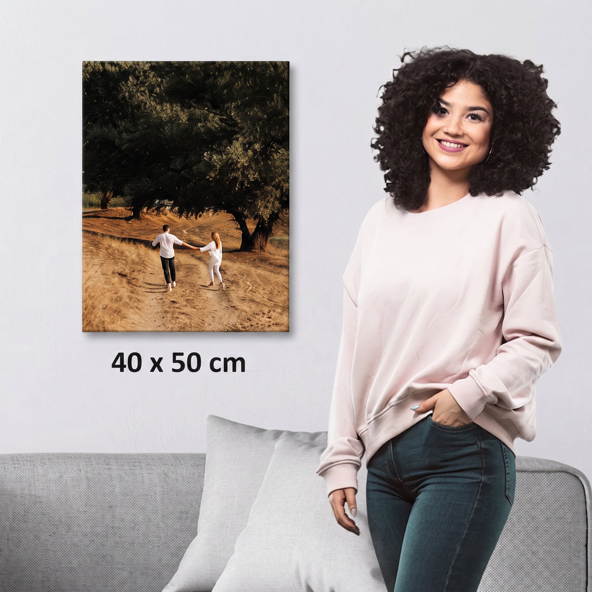 A woman stands beside a gray sofa wearing a light pink sweatshirt and dark jeans, while a framed-style wall art print showing a couple walking in an olive grove is displayed above her. The print includes the text '40 x 50 cm' to indicate size.