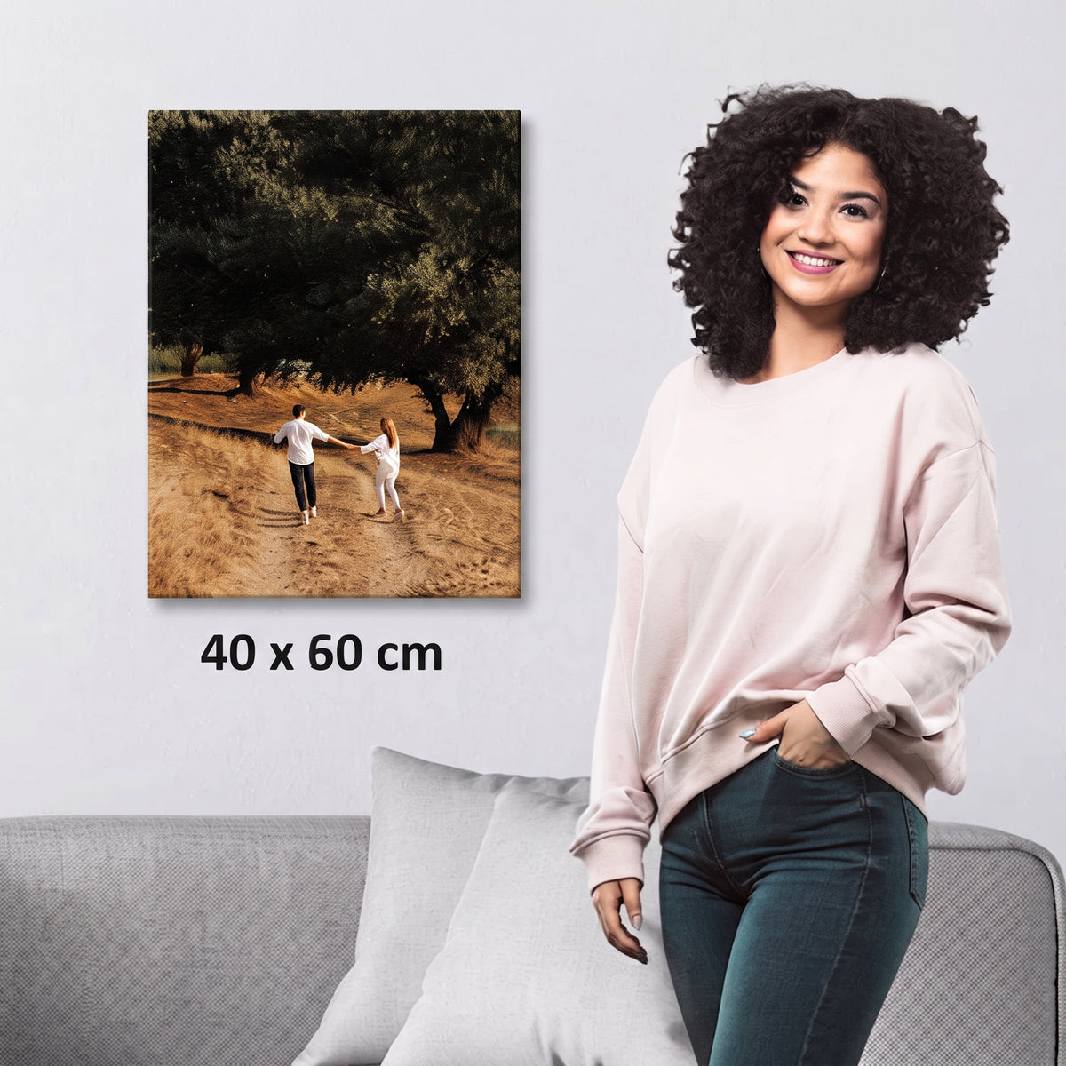 A woman stands beside a wall-mounted canvas print showing a couple walking in a sunlit tree-lined path. The print is labeled 40 x 60 cm, and the scene includes a sofa and pillows in a styled interior.