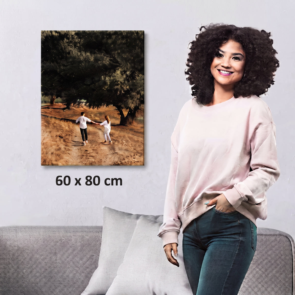 A woman stands beside a gray sofa in a living room setting, with a framed-style wall art print showing an outdoor scene of two people walking under trees. The print includes the text '60 x 80 cm' below it.