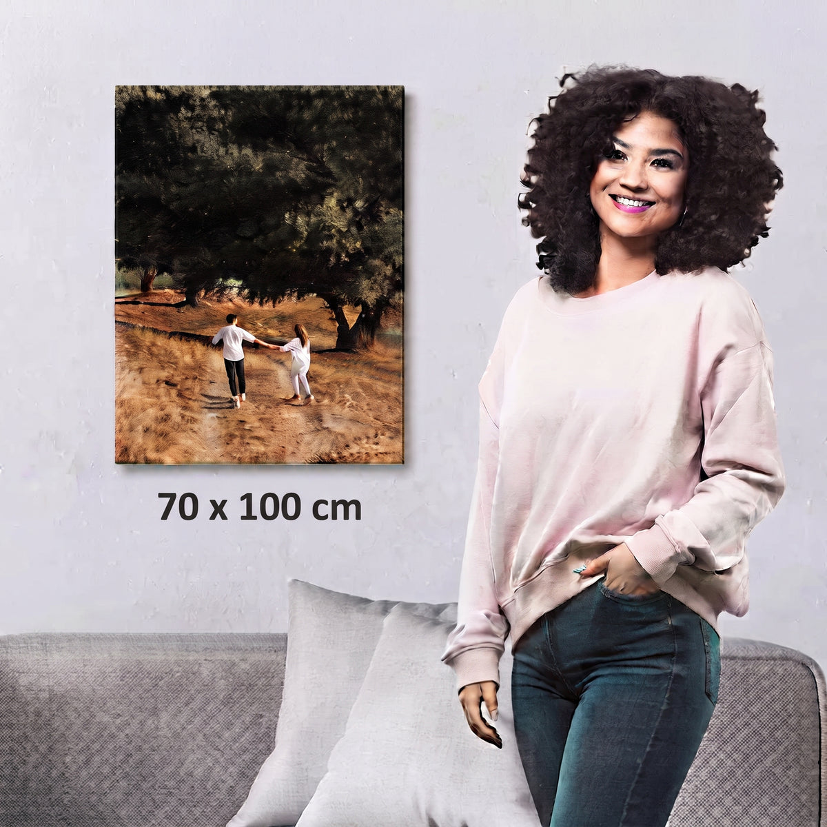 A woman stands beside a framed custom canvas print of two children playing in a grassy field under trees, with the text '70 x 100 cm' below the artwork. The canvas is shown hanging on a light gray wall above a gray sofa with pillows.
